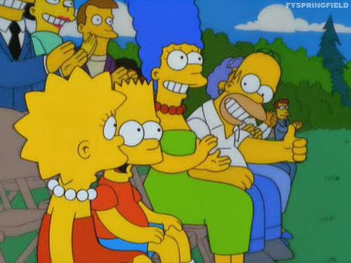 The simpsons family sitting on bleachers at a sports game. Homer is smiling a big smile and giving Lisa a thumbs up.
