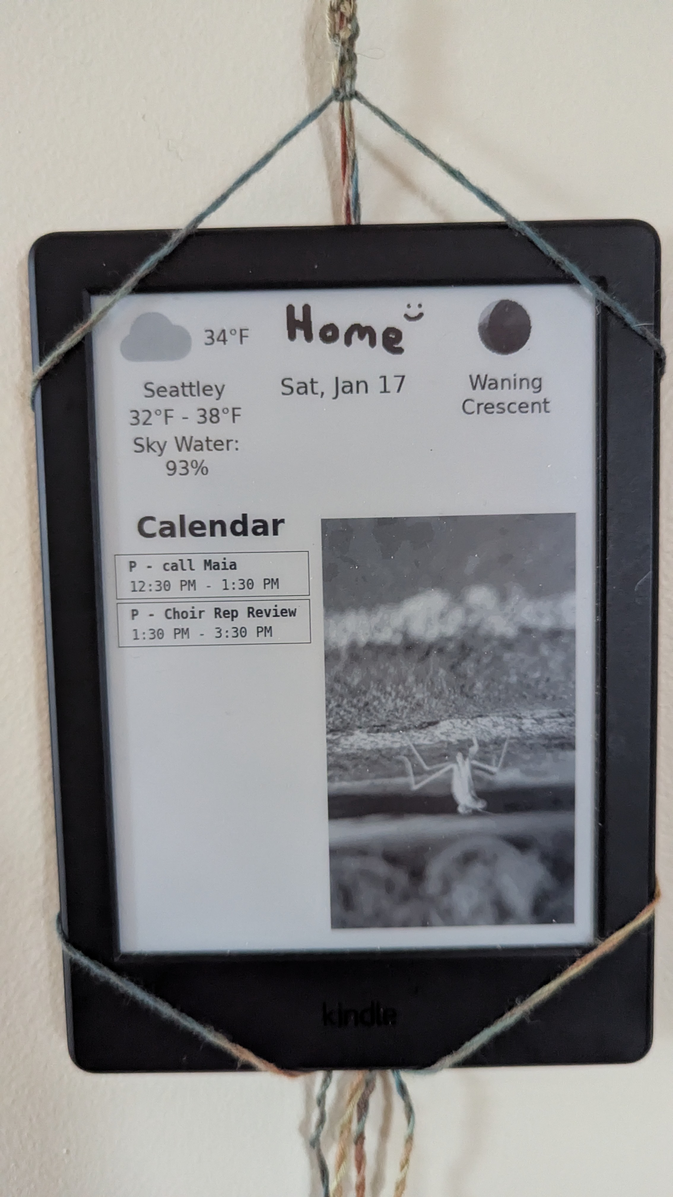 A kindle hanging on the wall from a macramé holder. The kindle is displaying a dashboard display. The display has two rows. The first row has the current weather on the left (34 degrees farenheight and Seattle-y), a pixel art drawing of the word Home and a smiley face in the middle, the date (Jan 17) below it, and the current moon phase to the right. The second row has a Calendar widget, with a list of calendar entries, including reminders to call a friend and to go to choir. On the right side is a greyscale image of a grasshopper climbing a wall that takes up the rest of the space on the display.