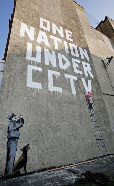 A bansky mural from 2007, depicting a child being forced to paint the slogan "One Nation Under CCTV" by a cop and his dog. A real CCTV camera is mounted just to the right of the mural.