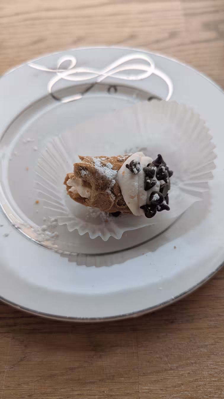 A half eaten plain cannoli from Holy Cannoli! It's sitting in a little paper wrapper on top of a small plate, with powdered sugar dusting everything. The cannoli has white frosting and little chocolate chips on one end.