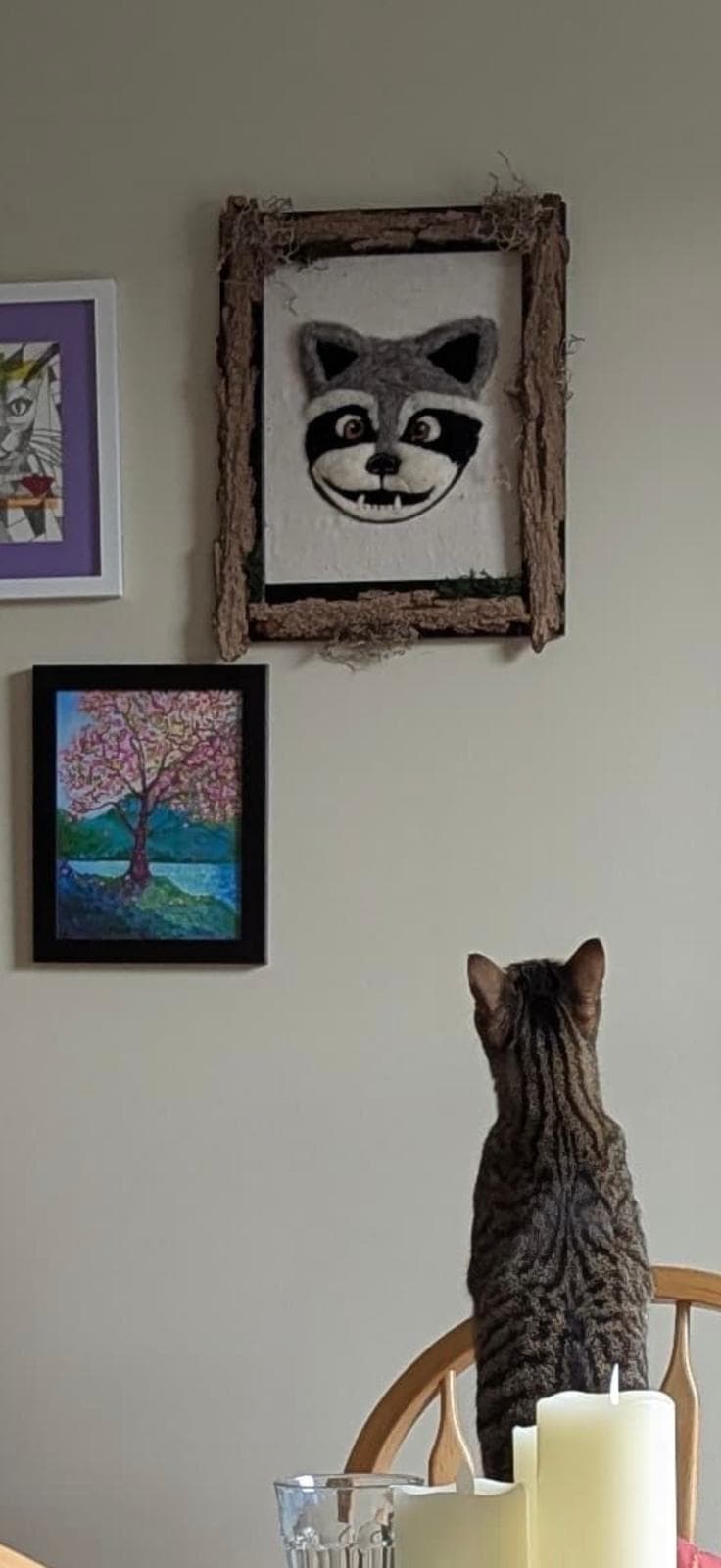 Artie the cat stands on a chair to stare up at the new art hung on the walls. The art is a framed canvas containing a needle-felted raccoon face, grinning at the viewer. The frame is decorated with glued-on pieces of tree bark and moss.